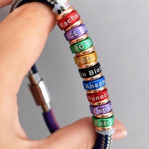 May include: A colourful bracelet featuring multiple rings, each inscribed with a different word or name. The rings are in various colours, including red, purple, green, and blue. The bracelet is held by a hand.