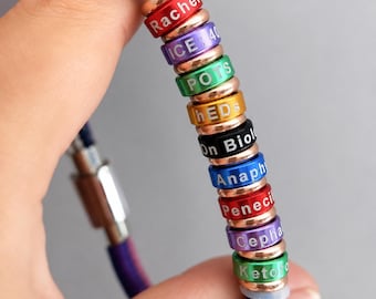 Puede incluir: Una pulsera multicolor con varios anillos, cada uno mostrando una palabra o nombre diferente. Los anillos son de varios colores, incluyendo rojo, morado, verde y azul. La pulsera es sostenida por una mano.