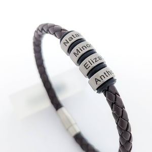 Op de afbeelding: Een bruin leren armband met vier zilveren kralen. Elke kraal is gegraveerd met een naam: "Natal", "Minor", "Eliza" en "Antho".