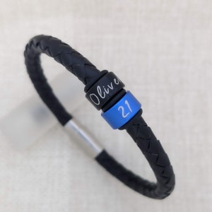 May include: Black leather bracelet with a silver slider and two engraved metal beads. One bead says "Oliver" and the other bead says "21".