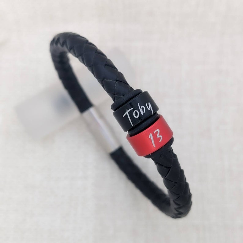 May include: Black leather bracelet with a black and red bead. The bead is engraved with the name "Toby" and the number "13".