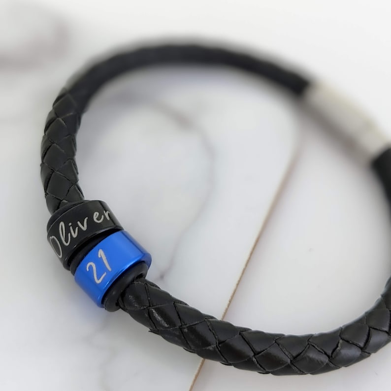 May include: Black leather bracelet with a blue and black bead engraved with the name "Oliver" and the number "21".