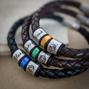 May include: Three leather bracelets with personalized metal beads. The beads are engraved with the names "Blake", "Gabriel", and "Noah". Each bead has a colored ring and a small metal charm.