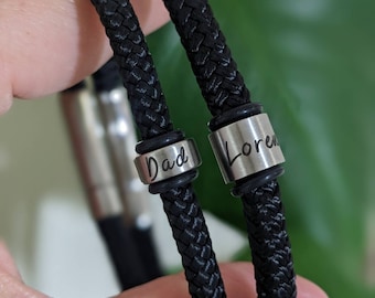 Puede incluir: Dos pulseras trenzadas negras con cuentas de metal plateado. Las cuentas están grabadas con las palabras "Dad" y "Loren".