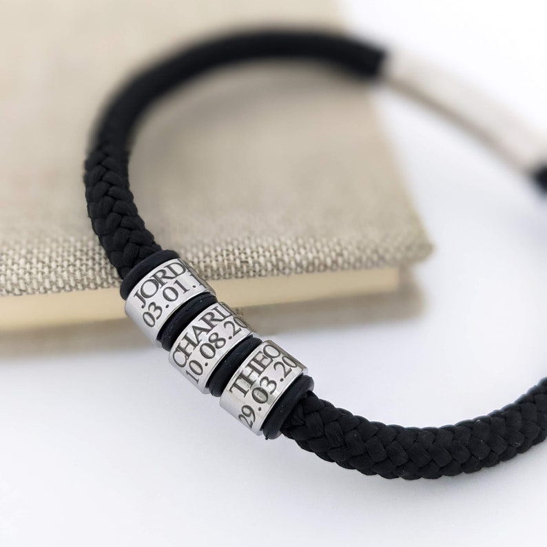 K&ouml;nnte beinhalten: Schwarz geflochtenes Armband mit drei silbernen Perlen, die mit Namen und Daten graviert sind. Das Armband hat eine dicke, gewebte Textur und einen Verschluss. Die Perlen sind mit den Namen "JORD", "CHARLI" und "THEO" sowie Daten graviert.
