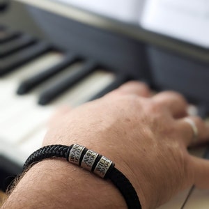 K&ouml;nnte beinhalten: Ein schwarzes geflochtenes Armband mit silbernen Perlen, die jeweils mit einem Namen und Datum graviert sind. Das Armband wird am Handgelenk getragen, mit einer Klaviatur im Hintergrund. Die Perlen lesen: BRAND 06.02.2, AMEL 13.08.3, WILL 29.04.