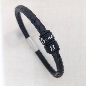 May include: Black leather bracelet with a silver clasp and two black metal beads. The beads are engraved with the name "Gann" and the number "13".