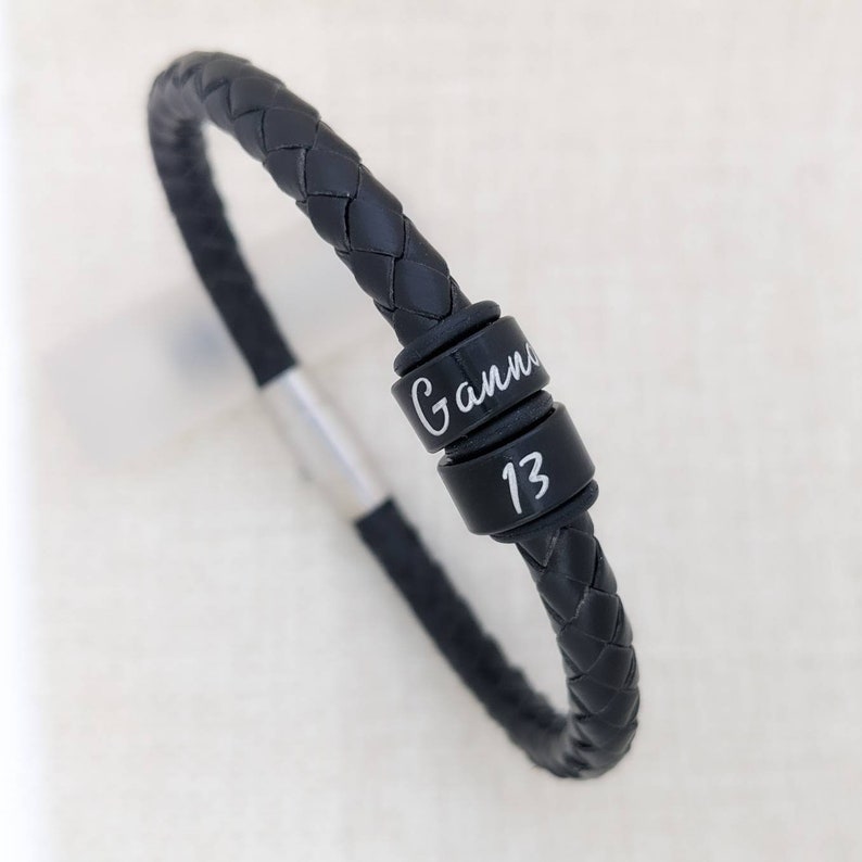 May include: Black leather bracelet with a silver clasp and two black metal beads. The beads are engraved with the name "Gann" and the number "13".