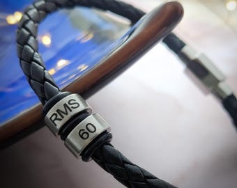 May include: Black braided leather bracelet with silver-tone metal accents. The bracelet features the text "RMS" and the number "60" on the metal beads. The bracelet is shown on a wooden surface.