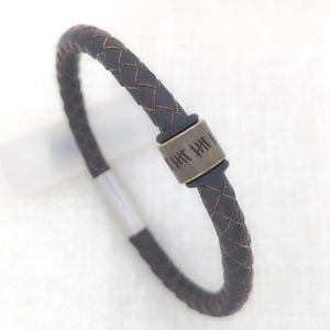 May include: A dark brown braided leather bracelet featuring a cylindrical metal bead with etched tally mark designs. The bracelet includes a smooth silver-toned metal clasp mechanism that secures the braided cord ends together.
