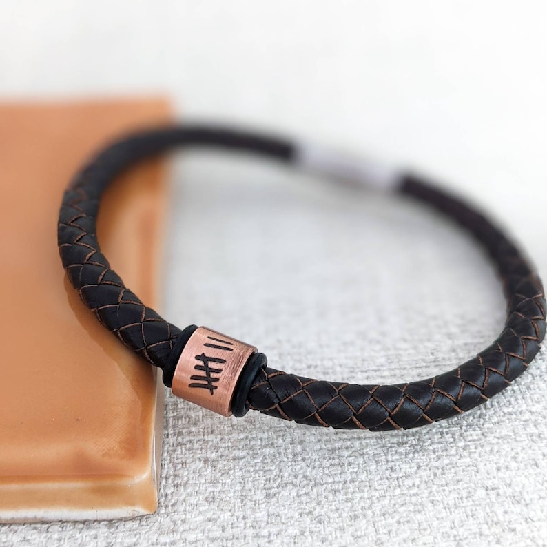Pu&ograve; includere: Un braccialetto in pelle nera con un perno in rame inciso con numeri romani. Il braccialetto &egrave; intrecciato e ha un design semplice.