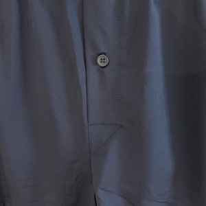 May include: A pair of solid dark navy blue boxer shorts featuring an elasticized waistband and a central front fly opening secured by a single matching dark navy blue button. The fabric has a smooth, matte finish with visible vertical stitching.