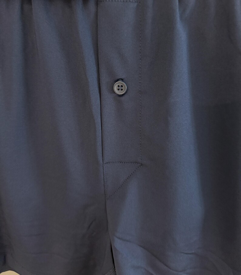 May include: A pair of solid dark navy blue boxer shorts featuring an elasticized waistband and a central front fly opening secured by a single matching dark navy blue button. The fabric has a smooth, matte finish with visible vertical stitching.
