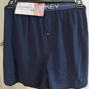 May include: Navy blue boxer shorts featuring an elastic waistband with the Jockey brand name printed in white. The garment includes a single front button closure and a product tag attached to the waistband that reads Jockey Eco-Friendly Microfiber.
