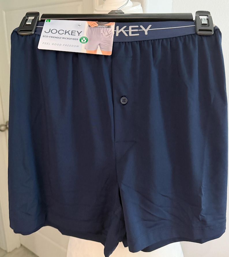 May include: Navy blue boxer shorts featuring an elastic waistband with the Jockey brand name printed in white. The garment includes a single front button closure and a product tag attached to the waistband that reads Jockey Eco-Friendly Microfiber.