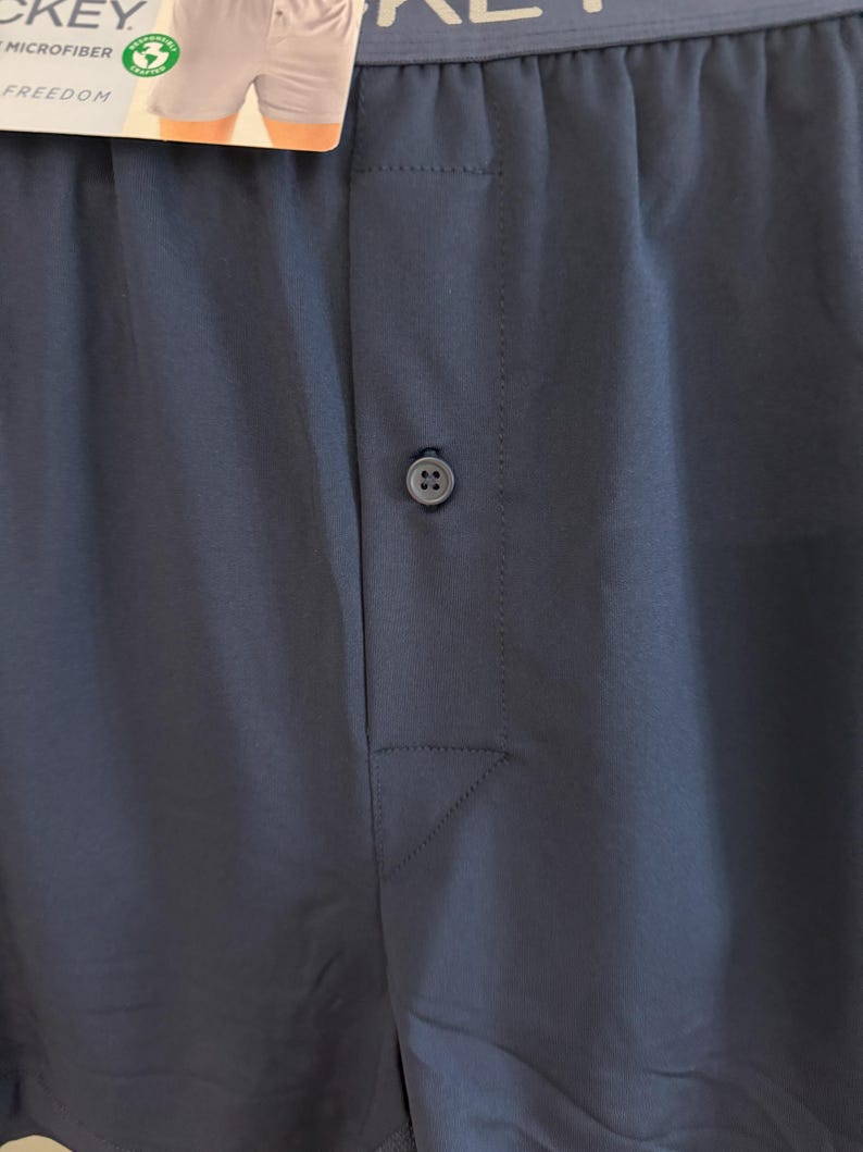 May include: A pair of dark blue boxer shorts featuring a central button closure on the fly. The waistband is elasticized and displays the text JOCKEY MICROFIBER FREEDOM. A product tag is attached to the top, showing a partial view of the garment.