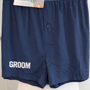 May include: Navy blue boxer shorts featuring a single button fly and the word GROOM printed in white on the lower left leg. The waistband is elastic with the Jockey brand name in white text and includes an attached product tag.