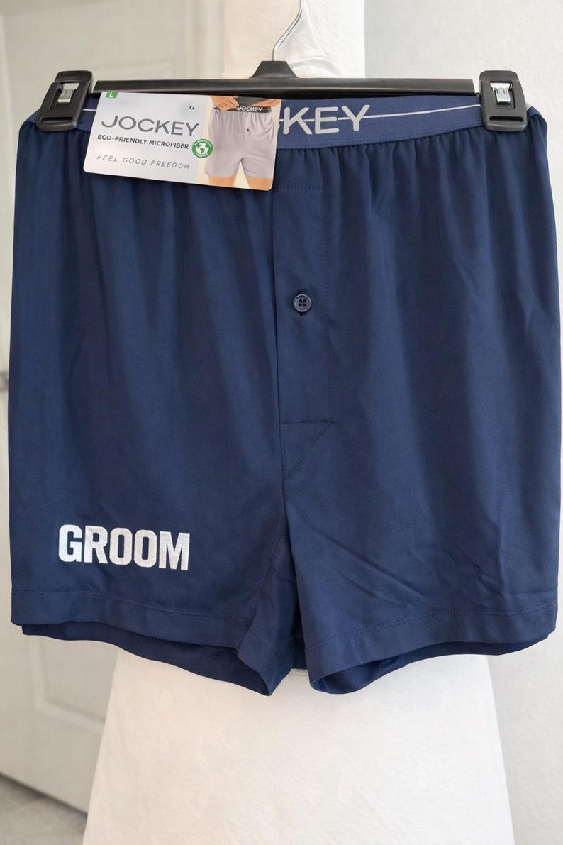 May include: Navy blue boxer shorts featuring a single button fly and the word GROOM printed in white on the lower left leg. The waistband is elastic with the Jockey brand name in white text and includes an attached product tag.