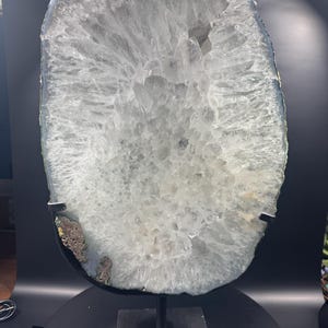 May include: A large, oval-shaped translucent crystal geode slice with a jagged, crystalline interior texture in shades of clear and white. The geode is mounted on a black metal stand with a square base, featuring a dark, rough outer edge.