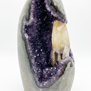 Pode incluir: Uma geodo de ametista vertical com um exterior cinzento liso e arredondado e um centro oco revestido com cristais roxos facetados. Uma única ponta de calcite ou quartzo amarelo pálido e translúcido ergue-se dentro da cavidade cristalina.