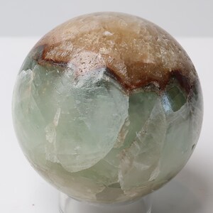 May include: A polished spherical mineral specimen featuring a translucent pale green base with a distinct band of opaque tan and dark brown crystalline material across the upper portion, showcasing natural internal fractures and varied mineral textures.