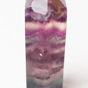 May include: A polished fluorite crystal tower featuring a hexagonal point and a smooth, multifaceted surface. The stone displays a gradient of translucent purple, deep violet, and subtle green bands with natural internal inclusions and fractures.
