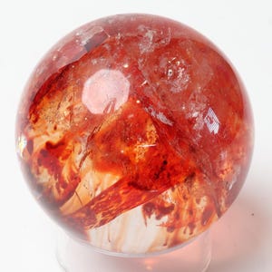 May include: A polished spherical crystal featuring deep red and orange inclusions suspended within a clear, translucent quartz base. The stone displays natural internal fractures and varied colour density, creating a complex, marbled appearance.