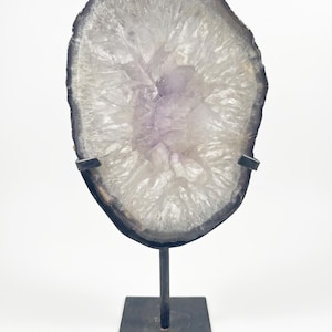 May include: A translucent, oval-shaped amethyst geode slice with a dark, jagged outer edge, mounted on a black metal stand. The crystal features a pale purple center that radiates outward into clear, crystalline formations with a textured surface.