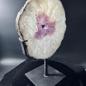 May include: A polished agate slice featuring a translucent white crystalline outer ring and a vibrant purple amethyst center, mounted vertically on a black metal stand with a square base, showcasing natural mineral textures and jagged edges.