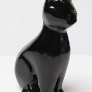 May include: A smooth, polished black cat figurine in a seated position. The sculpture features a glossy, reflective surface with a minimalist silhouette, pointed ears, and a rounded head, capturing the feline form in a solid, dark, opaque material.