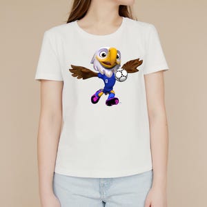 Puede incluir: Una camiseta blanca de manga corta con un gr&aacute;fico de una mascota de &aacute;guila de dibujos animados que viste un uniforme de f&uacute;tbol azul. El &aacute;guila aparece en movimiento, sosteniendo un bal&oacute;n de f&uacute;tbol blanco y negro con su ala y usando botas rosas.