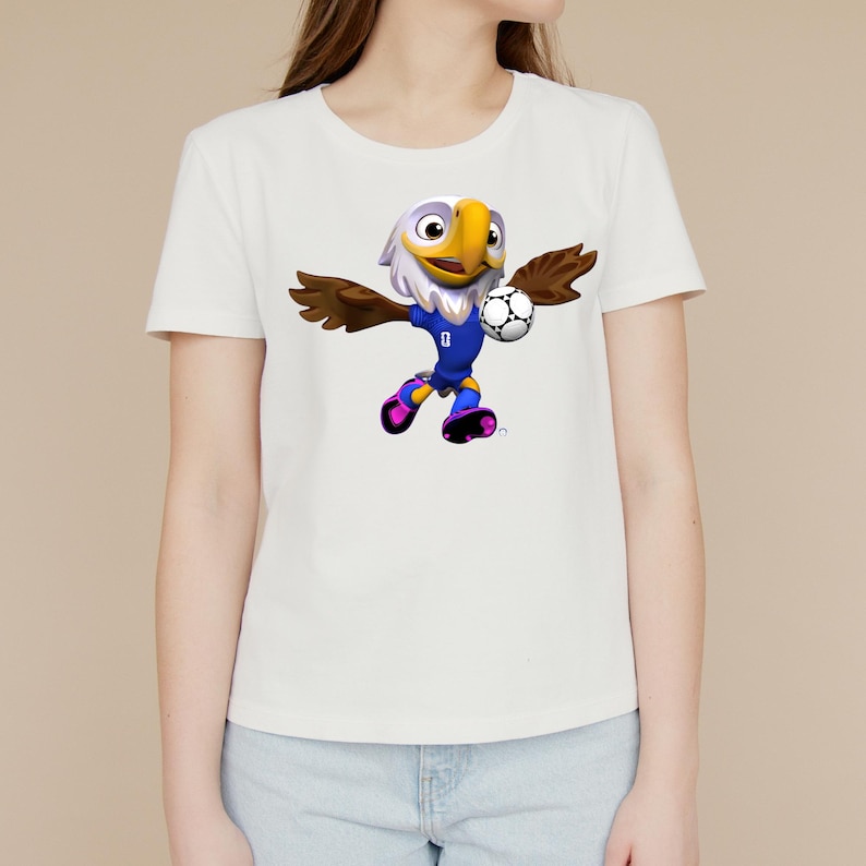 Puede incluir: Una camiseta blanca de manga corta con un gr&aacute;fico de una mascota de &aacute;guila de dibujos animados que viste un uniforme de f&uacute;tbol azul. El &aacute;guila aparece en movimiento, sosteniendo un bal&oacute;n de f&uacute;tbol blanco y negro con su ala y usando botas rosas.