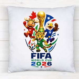 Puede incluir: Un coj&iacute;n cuadrado blanco con un gr&aacute;fico colorido del logotipo de la Copa Mundial de la FIFA 2026. El dise&ntilde;o incluye un trofeo dorado, un bal&oacute;n de f&uacute;tbol y tres mascotas con camisetas rojas, azules y verdes con el n&uacute;mero 26 y una hoja de arce.