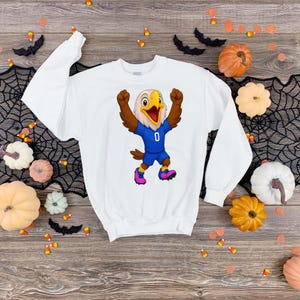 Puede incluir: Una sudadera blanca de manga larga con un gr&aacute;fico de un &aacute;guila de dibujos animados. El &aacute;guila tiene alas marrones, un pico amarillo y viste una camiseta azul con el n&uacute;mero cero en el pecho, pantalones cortos azules y zapatillas deportivas rosa.