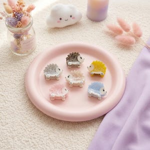 Puede incluir: Seis pequeñas pinzas para el cabello con forma de erizo en varios colores, incluyendo marrón, amarillo, azul claro, rosa, blanco y gris, dispuestas sobre un plato redondo y liso de color rosa claro. Cada pinza muestra un diseño de erizo detallado.
