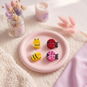 Puede incluir: Cuatro pequeñas pinzas para el pelo dispuestas sobre un plato rosa redondo. Dos pinzas son abejas amarillas con rayas negras y alas transparentes, y dos son mariquitas, una roja y otra rosa, ambas con puntos negros y mecanismos de agarre.