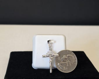 May include: A silver-toned crucifix pendant featuring a central figure and a small fan-shaped arrangement of clear stones above the crossbar. The pendant is displayed next to a United States quarter coin for scale, showing its relative size.