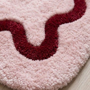 May include: A soft, tufted rug featuring a light pink base with a prominent, deep burgundy wavy line running horizontally across the surface. The texture is thick and plush, creating a dense pile appearance with a distinct, curved pattern.