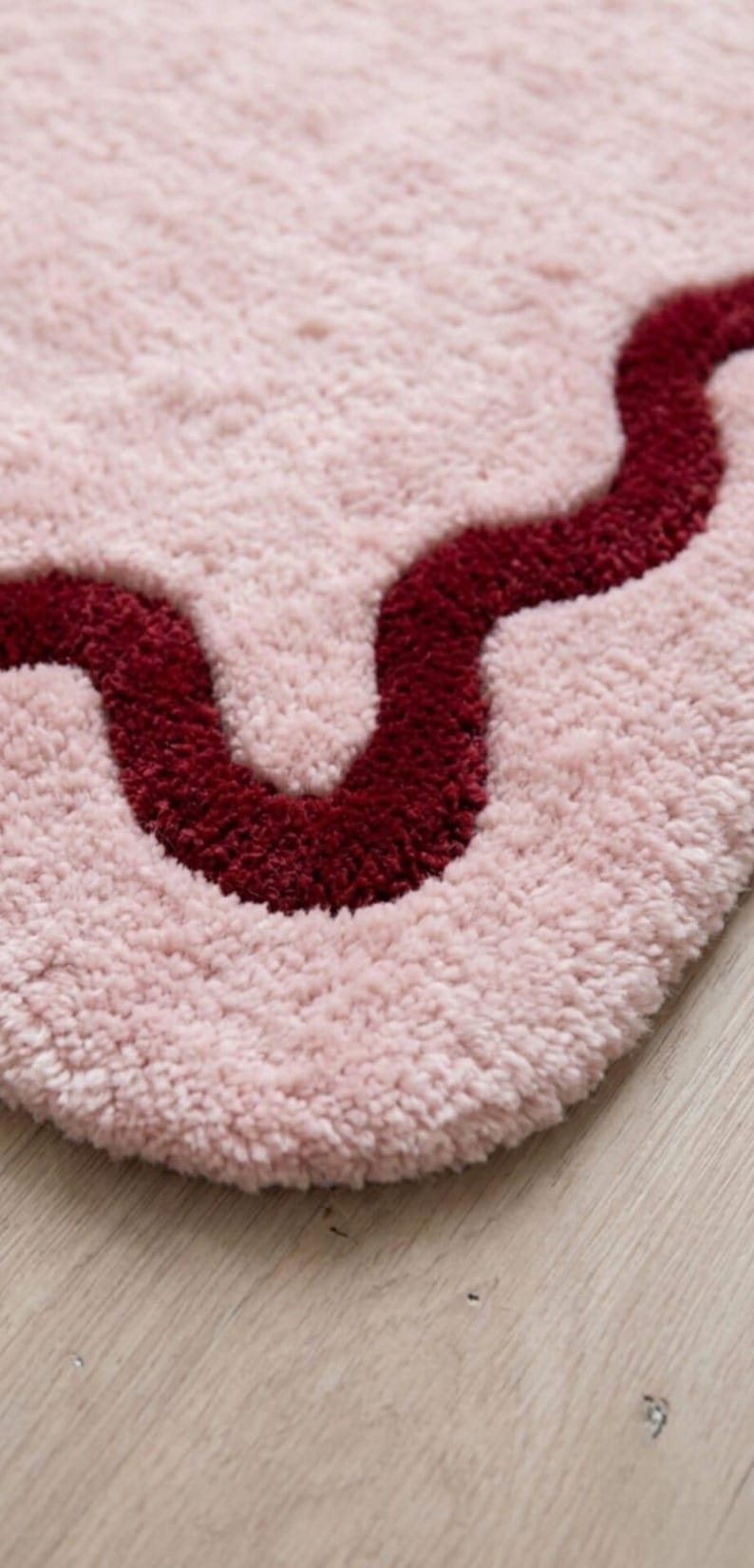 May include: A soft, tufted rug featuring a light pink base with a prominent, deep burgundy wavy line running horizontally across the surface. The texture is thick and plush, creating a dense pile appearance with a distinct, curved pattern.