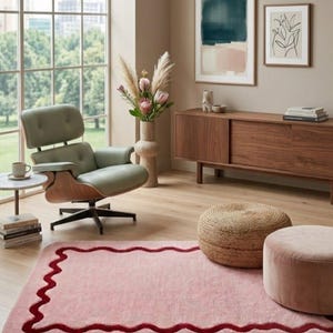 May include: A rectangular pink area rug featuring a dark red wavy border design. The rug is placed on a light wood floor in a room containing a sage green lounge chair, a wooden sideboard, two round ottomans, and wall art with the text ZINDAGI SE.