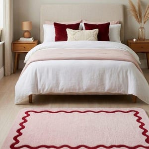 May include: A rectangular area rug featuring a soft light pink surface with a dark red scalloped border. The rug has rounded corners and a plush texture, positioned on a light-colored wooden floor in front of a bed with white and pink bedding.