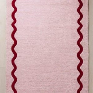 May include: A rectangular light pink rug featuring a deep red wavy border that follows the perimeter of the mat. The rug has a soft, plush texture with rounded corners and a consistent, solid color fill within the scalloped red line design.