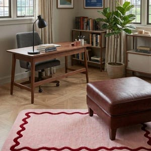 May include: A rectangular area rug featuring a soft pink center and a dark red wavy border. The rug is placed on a light-colored wooden floor with a herringbone pattern, positioned in front of a brown leather ottoman in a home office setting.