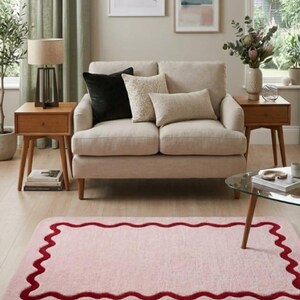 May include: A rectangular area rug featuring a soft light pink base color with a dark red scalloped border design. The rug has rounded corners and a plush, textured surface, positioned on a light-colored wooden floor in a living room setting.