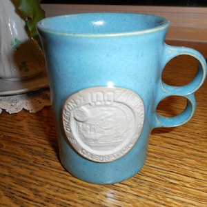 May include: A blue ceramic mug with two handles. The mug has a white circular design with the text "Oregon 100th Anniversary Celebration".