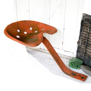 May include: An orange metal tractor seat with a rusty finish and a curved metal support. The seat has several holes in it.