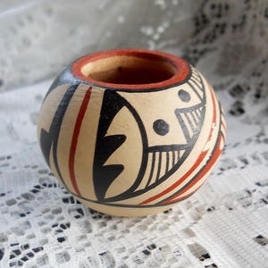 May include: A small, hand-painted ceramic pot with a geometric design in black, red, and white. The pot has a rounded shape and a smooth, matte finish.