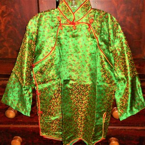 K&ouml;nnte beinhalten: Eine gr&uuml;ne Seidenjacke im chinesischen Stil mit goldenen und roten Akzenten. Die Jacke hat einen Mandarinkragen und einen Knopfverschluss. Der Stoff ist mit kleinen Blumenmustern gemustert.