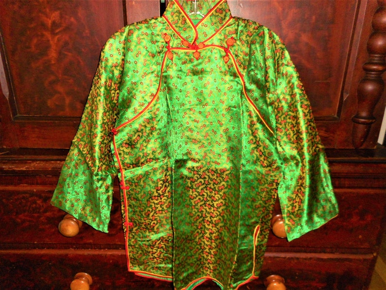 K&ouml;nnte beinhalten: Eine gr&uuml;ne Seidenjacke im chinesischen Stil mit goldenen und roten Akzenten. Die Jacke hat einen Mandarinkragen und einen Knopfverschluss. Der Stoff ist mit kleinen Blumenmustern gemustert.