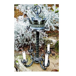 May include: A black and green metal chandelier with a crown-shaped top and four candle-style light bulbs. The chandelier is in a garden setting.
